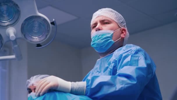 男性外科医の肖像画 真剣に前を見てマスクと保護制服の医者 診療所での手術中に外科医と看護師 — ストック動画