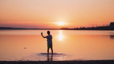 Akşamları bir çocuğun arka görüntüsü. Çocuk gün batımında suda duruyor ve nehre kağıt uçak fırlatıyor. Yavaş çekim.