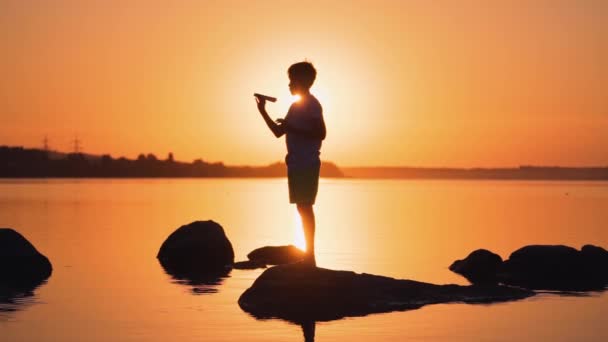 日落时带着纸飞机的男孩的轮廓 小孩站在橙色河畔的一块石头上 晚霞的光芒照耀着一个男孩 — 图库视频影像