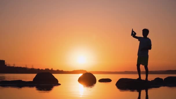 一个夏天玩折纸飞机的男孩日落时 一个小孩在桔子湖中的小岩石上的轮廓 慢动作 — 图库视频影像