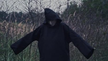 Kapüşonlu, koyu renk kostümlü korkunç adam. Korkunç bir hayaletin portresi. Kolları doğa arka planında. Cadılar Bayramı konsepti.