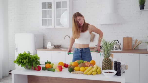 苗条的女人在厨房里测量她的腰围 穿着牛仔裤的快乐女孩站在桌旁 满屋子都是新鲜的水果和蔬菜 小姐瘦了 — 图库视频影像