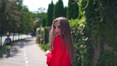 Yazın açık havada çekici bir kız. Tutkulu kırmızı elbiseli uzun saçlı kadın kameraya bakıyor ve caddede gülümsüyor. Yavaş çekim.