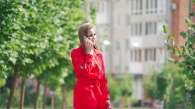 Kırmızı elbiseli güzel, uzun saçlı bir kadın. Parlak bir yaz gününde şehrin arka planında cep telefonuyla konuşan çekici bir bayan. Yavaş çekim.