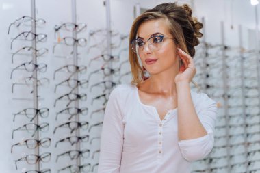 Mutlu bayan müşteri ya da gözlükçü optik dükkanında arka planda gözlük setiyle ayakta duruyor. Gözlüklü kadın. Görme düzeltmesi. Gözlüklü kız. Gözlüklü bir kadının portresi. Yakın çekim..
