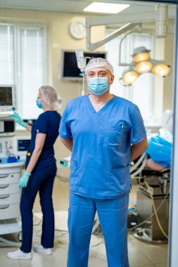 Maskeli bir cerrah kameraya bakıyor. Ameliyathanedeki doktor. Ekip arka planda çalışıyor..