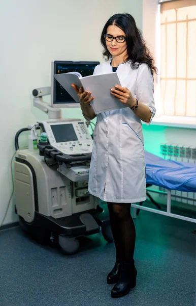 Female sonographer standing near ultrasound machine in clinic. Reading documents confidently.