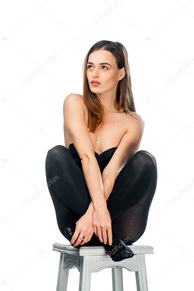Pretty woman posing sitting in black top and leggins on a chair over white background. Woman with long dark hair sits with crossed hands and crossed legs.