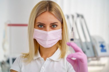 Elindeki pembe eldivenli tıbbi şırınga. Arka plan bulanık. Tıbbi maskeli doktor kadın. Enjeksiyon kavramı.