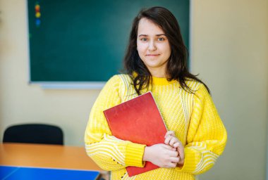 Güzel gülümseyen kız kitap tutuyor. Okula gidiyorum. Portre, sınıf arkaplanı. Öğrenci bir kitaba sarılıyor. Yaşam tarzı, ilgi, hobi, boş zaman, boş zaman.