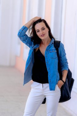 Sırt çantalı sıradan bir kadın. Mavi gömlekli ve beyaz kotlu genç kadın manzarası. Kameraya bakıyorum..