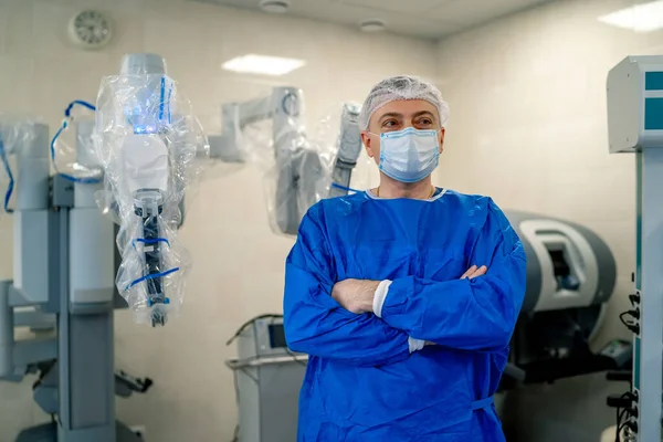 Medical specialist standing cross hands near modern surgery equipment. Doctor in hospital wearing scrubs and mask.