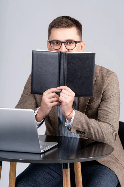 Sales manager has hidden face behind notebook. Laptop on table. Working in distance.