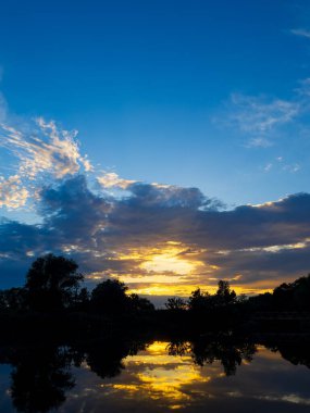 Gökyüzü birkaç bulutla mavidir ve güneş batıyor. Su sakin ve gökyüzünü ve ağaçları yansıtıyor.