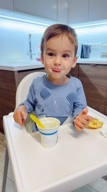 Genç bir çocuk yüksek bir sandalyede oturmuş yoğurt yiyor. Ağzında bir kaşık var ve bir parça ekmek tutuyor..