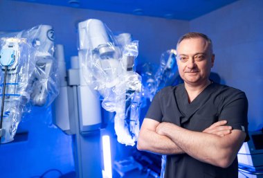 Cerrah robot ameliyathanede duruyor. Bir cerrah, gelişmiş robotik cerrahi ekipman ve mavi ışıklandırmanın yer aldığı modern bir ameliyathanede güvenle poz verir..