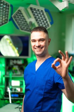 Cerrah güven yayar. Mavi önlüklü bir cerrah iyi aydınlatılmış bir ameliyathanede duruyor, gülümsüyor ve ameliyat sırasında iyi bir jest yapıyor..