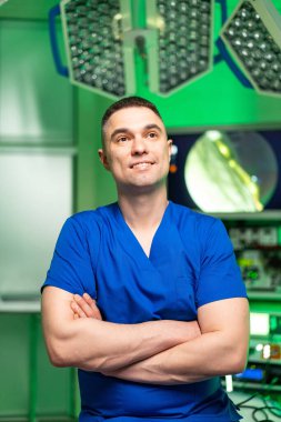 Mavi önlüklü, gülümseyen bir cerrah. Cerrah, modern ve parlak bir ameliyathanede kollarını kavuşturarak tıbbi bir prosedüre hazırlanıyor..