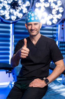 Gülümseyen cerrah ameliyathanede onay verdi. Kendine güvenen bir cerrah başarılı bir ameliyata hazır, iyi aydınlatılmış bir ameliyathanede baş parmağını paylaşıyor..