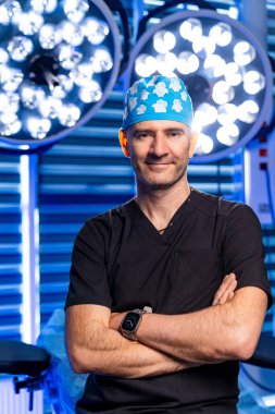 Cerrah ameliyata hazır. Cerrah önlüklü, kollarını kavuşturmuş ameliyat odasının ışıklarıyla parlak bir şekilde duruyor..