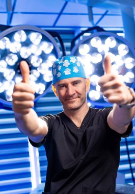 Cerrah başarılı bir operasyonu kutluyor. Ameliyat önlüğü ve cerrahi şapkalı cerrah arka planda parlak cerrahi ışıkları olan bir ameliyathanede baş parmak gösteriyor..