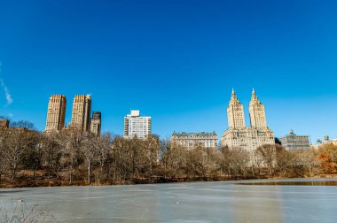 Central Park 'ta kış. New York 'taki Central Park' ta açık mavi bir gökyüzü sergilenirken, binalar kışın dingin gölü çerçeveliyor..