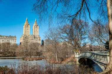 Central Park 'ta kış gökdelenleri. Central Park 'ta çıplak ağaçlar ve açık mavi gökyüzüne karşı görünen ikonik binaları olan bir köprü sergileniyor..