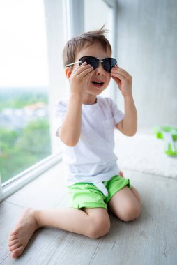 Çocuk içeride güneş gözlükleriyle oynuyor. Parlak giyimli bir çocuk, güneşli bir günün keyfini çıkarırken yerde oturur ve güneş gözlüğünü ayarlar..