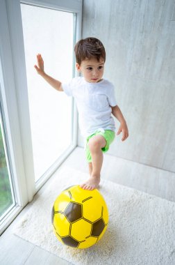 Çocuk içeride futbol oynuyor. Genç bir çocuk pencereye yaslanırken parlak sarı bir futbol topunun üzerinde dengede duruyor. Güneş ışığı odayı dolduruyor..
