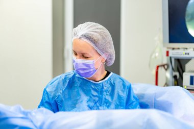 Ameliyat önlüğü giymiş bir cerrah. Mavi önlüklü ve maskeli sağlık çalışanı ameliyat sırasında steril bir ortamda cerrahi görevlere odaklandı..