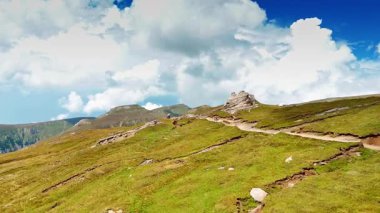 Yürüyüşçüler öğleyin yemyeşil tepeleri keşfederler. Yürüyüşçüler, parlak mavi bir gökyüzünün altında, gün ortasında kabarık bulutlarla dolu dolambaçlı yeşil tepelerde dolaşırlar..
