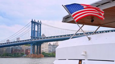 Manhattan Köprüsü 'nün tekneden görünüşü. Manhattan Köprüsü 'nün altında Amerikan bayrağını ve şehir siluetini gösteren bir tekne alacakaranlıkta sakince yelken açar..