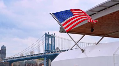 Bayraklı Brooklyn Köprüsü. Nehirde Amerikan bayrağı taşıyan bir tekne, Brooklyn Köprüsü bulutlu bir gökyüzünün altında..