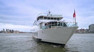 Şehir limanında lüks bir tekne geziyor. Şık bir tekne, şehir limanındaki dalgalı sularda, bulutlu bir gökyüzünün altında yol alır. Şehir siluetini gözler önüne serer..