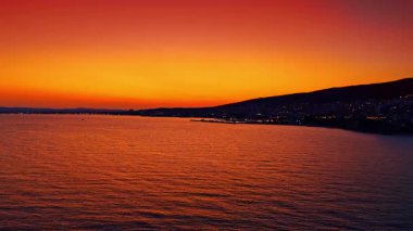 Sakin dalgalarda canlı gün batımı. Güneş sakin denizin üzerinde batarken, kıyı şeridini aydınlatan canlı turuncu ve mor renkler gökyüzünü dolduruyor..