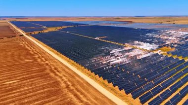 Açık gökyüzü altında geniş bir güneş çiftliği. Geniş güneş panelleri tarlalar boyunca uzanıyor, güneşli bir günde güneşleniyorlar, yenilenebilir enerji çabalarını vurguluyorlar.
