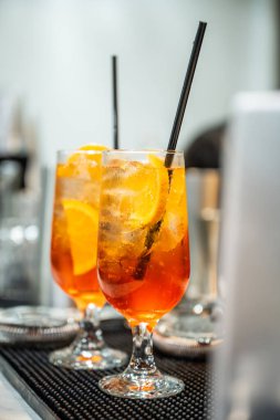 Barda servis edilen yaz içkilerini tazeliyorum. Portakal dilimleriyle süslenmiş parlak turuncu bir içecekle dolu iki bardak bir bar tezgahına oturur..