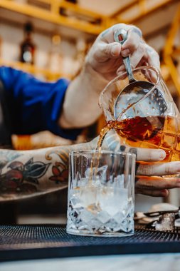 Denver, ABD, 14 Ağustos 2024: Canlı bir barda kokteyl hazırlamak. Bir barmen ustalıkla buzun üzerine altın bir içki döker. Yoğun bir gecede kokteyl sanatını sergiler..