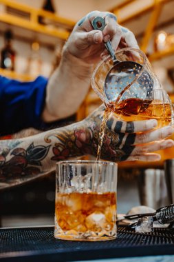 Denver, ABD, 14 Ağustos 2024: Yetenekli barmen mükemmel bir kokteyl hazırlıyor. Yetenekli bir barmen buzla dolu bir bardağa iyi hazırlanmış bir kokteyl koyar. Karmaşık dövmeler kollarını süslüyor..