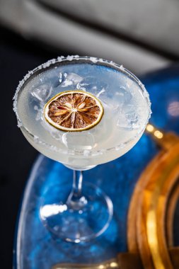 Şık bir bardakta servis edilen zarif bir kokteyl. Kurutulmuş kireçle süslenmiş ferahlatıcı bir kokteyl mavi bir masada oturur ve eşsiz sunumuyla dikkatleri üzerine çeker..
