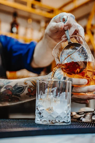 Denver, ABD, 14 Ağustos 2024: Canlı bir barda kokteyl hazırlamak. Bir barmen ustalıkla buzun üzerine altın bir içki döker. Yoğun bir gecede kokteyl sanatını sergiler..