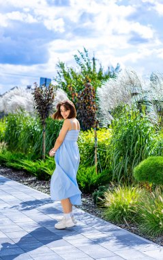 Genç bir kadın, yaz kıyafetleriyle bahçede dolaşmaktan hoşlanıyor. Açık renk elbiseli genç bir kadın açık mavi gökyüzünün altında yemyeşil bir bahçede kıvrılır..