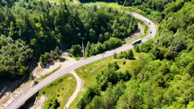 Parlak bir günde yeşil tepelerde kıvrılan kıvrımlı bir yol. Araçlar, açık mavi gökyüzünün altında yemyeşil ağaçlar ve yemyeşil ağaçlarla çevrili virajlı bir yolda seyahat ederler..