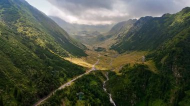 Dağ vadisi manzarası kıvrımlı yol ve akarsu. Sakin bir dağ vadisinde dolambaçlı bir yol, çimenli düzlükler ve yemyeşil sularda akan yumuşak bir akarsu bulunur..