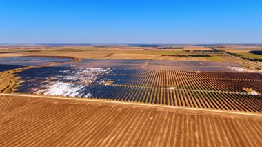 Kuru tarım arazisi boyunca güneş enerjisi santrali. Güneş panelleri parlak gökyüzünün altında açık tarım arazilerini kaplayan büyük bloklar halinde sıralanmış..