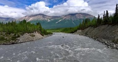İnsansız hava aracı görüntülerinden gri kirli suyla çalkantılı bir dağ nehri. Arka planda kabarık bulutlu kayalara çarpan. Alaska, ABD.