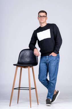 Genç adam bir stüdyoda kendine güvenen bir sandalyeyle poz veriyor. Genç bir adam gülümserken modern bir sandalyenin yanında duruyor. Basit bir stüdyo ortamında günlük kıyafetler giyiyor..