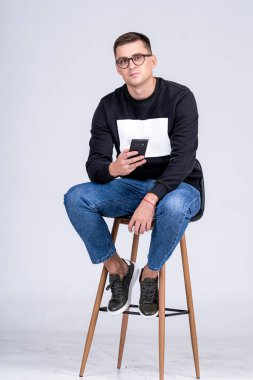 Genç adam taburede oturuyor ve akıllı telefon kullanıyor. Genç bir adam uzun bir taburede oturur, akıllı telefonuna, günlük kıyafetine, stüdyo geçmişine ve hafif ışıklandırmasına odaklanır..