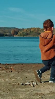Enerjik erkek bebek nehir kıyısında taş topluyor. Sıcak ceketli çocuk suya taş atıyor. Dikey video