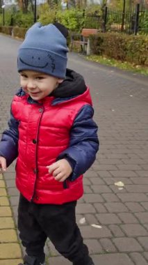 Şapkalı ve ceketli sevimli çocuk parkta. Aktif çocuk mutlu bir şekilde zıplıyor ve gülümsüyor. Dikey video